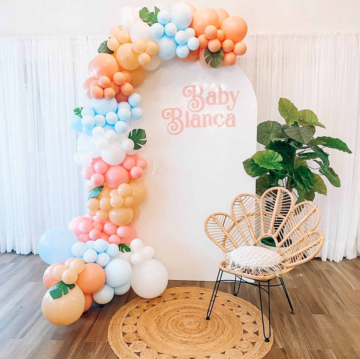 Baby shower backdrop in white and pink letters that read “baby Bianca” in a tropical font. Rattan boho flower accent chair, boho rug and balloons in white, blue, pink and peach colors with monstera leaves attached to them. 